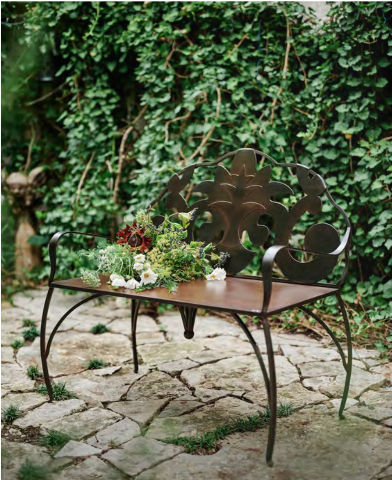 A hand-forged Jan Barboglio iron bench with scrollwork back, set on a stone courtyard amid ivy | Cayen Home, Carmel-by-the-Sea